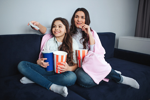 Beautiful brunette caucasian mother and daughter sit together in room. They watch movie. People hold popcorn baskets and cola cup. Adult has remote controller. Calm and concentrated