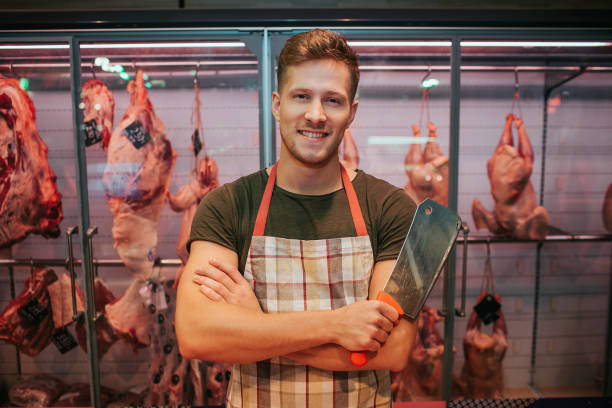 jeune homme se tiennent au plateau de viande dans l'épicerie. il tient le couteau de boucher à la main et pose. les animaux se braient derrière. travailleur masculin positif sourire. - butcher butchers shop meat store photos et images de collection