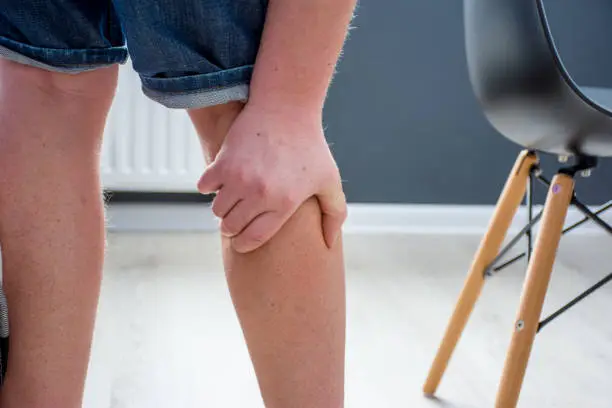 Photo of Human, who had been elevated from chair, holding his palm over calf or gastrocnemius muscle, which grabbed cramp with severe pain. Concept images illustrating human reaction to pain symptom in calf