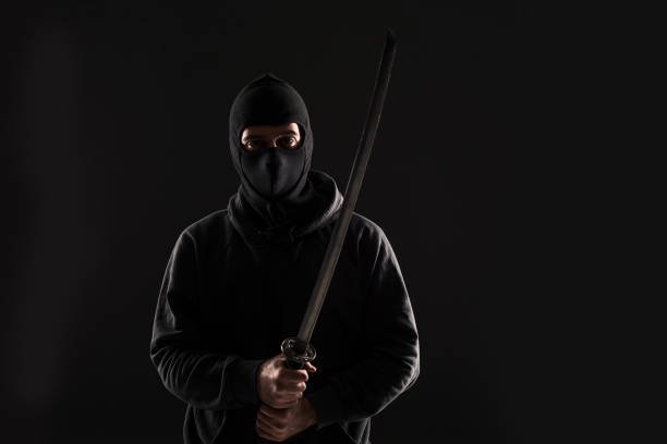 istock Man with balaclava and katana sword on black background 1136608645