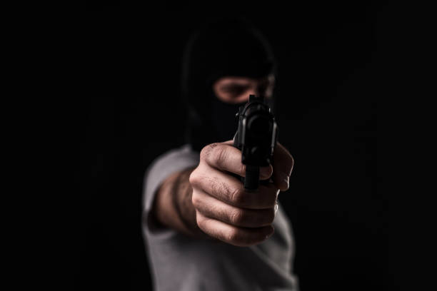 istock Masked robber with gun aiming into the camera against a black background 1136608662