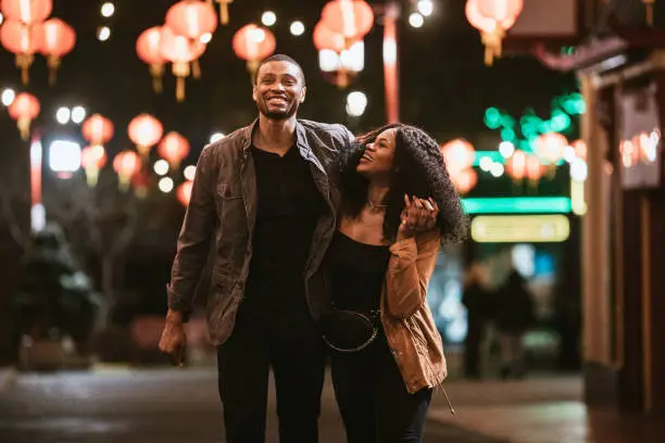 A smiling African American man and woman walk arm in arm on the streets of Chinatown in L.A. California on a warm evening, exploring the cities night life.  Bright traditional lanterns illuminate the scene.