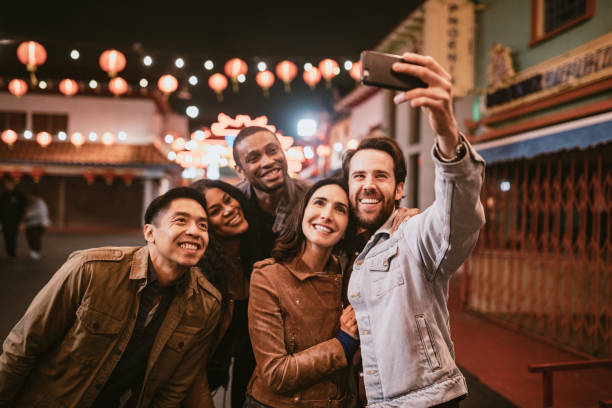 Friends Take Selfie in Chinatown Downtown Los Angeles At Night A diverse group of multiethnic friends walk the streets of Chinatown in L.A. California on a warm evening, exploring the cities night life.  Bright traditional lanterns illuminate the scene.  They take a smartphone self portrait to share on social media. financial district photos stock pictures, royalty-free photos & images