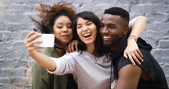 Shot of a group of friends taking a selfie together while posing outdoors against a grey brick wall