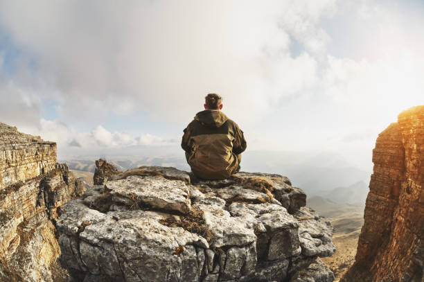 istock A young man sits on a rock surrounded by cliffs, located above the clouds in a great location, on a background of clouds, valleys, fields, mountains 1136619488