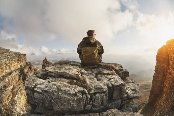 istock A young man sits on a rock surrounded by cliffs, located above the clouds in a great location, on a background of clouds, valleys, fields, mountains 1136619517
