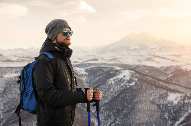 istock Young bearded hipster wearing a hat and sunglasses with Nordic walking sticks and a backpack moves in the mountains at sunset, against a background of sunset sky and volcano Elbrus 1136619713