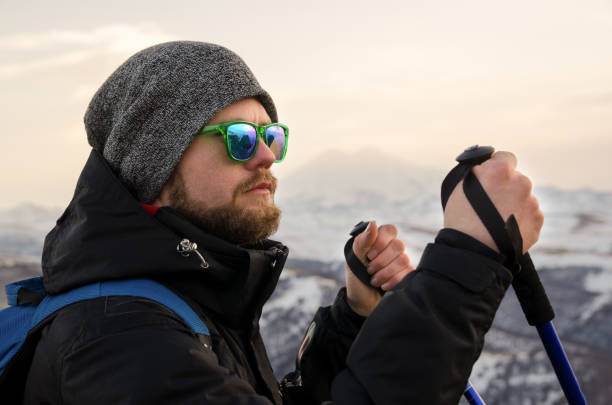 istock Young bearded hipster wearing a hat and sunglasses with Nordic walking sticks and a backpack moves in the mountains at sunset, against a background of sunset sky and volcano Elbrus 1136620032