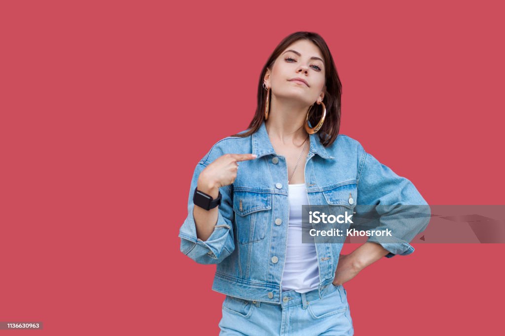 Portrait of proud beautiful brunette young woman with makeup in denim casual style standing, pointing herself and looking at camera with haughty face. Portrait of proud beautiful brunette young woman with makeup in denim casual style standing, pointing herself and looking at camera with haughty face. indoor studio shot, isolated on red background. Women Stock Photo