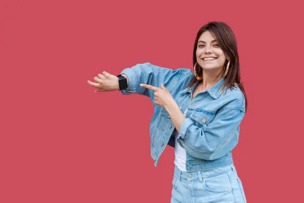 Photo of Portrait of happy beautiful brunette young woman with makeup in denim casual style standing showing and pointing at her smart watch with toothy smile