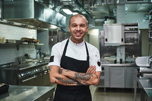 Me encanta mi trabajo alegre joven cocinero en delantal manteniendo los brazos tatuados cruzados y sonriendo mientras está de pie en una cocina de restaurante photo
