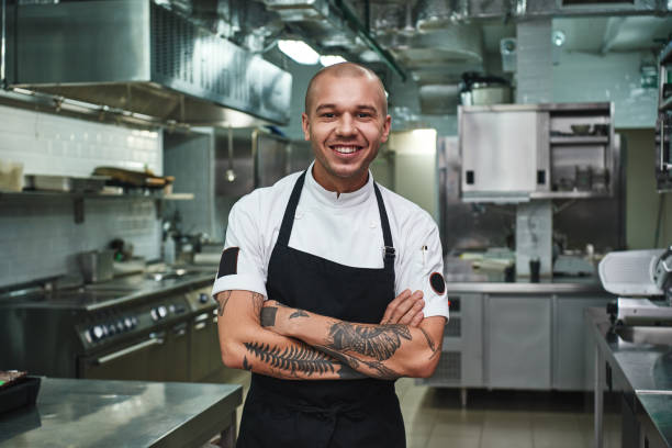 me encanta mi trabajo alegre joven cocinero en delantal manteniendo los brazos tatuados cruzados y sonriendo mientras está de pie en una cocina de restaurante - chef fotografías e imágenes de stock