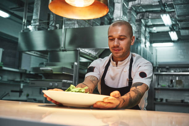 istock Ready to eat. Handsome smiling chef with tattoos on his arms, in black apron holding ready dish in modern restaurant kitchen. 1136638621