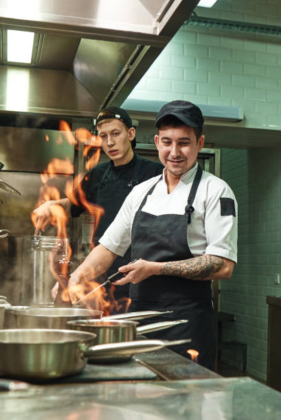 praca z ogniem. dwóch młodych i pozytywnych kucharzy uczy się, jak zrobić flambe na jedzenie w kuchni restauracji - pan frying pan fire fried zdjęcia i obrazy z banku zdjęć