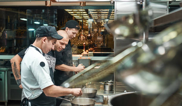 istock Culinary school. Strict young chef in apron looking carefully how his assistant cooking at restaurant kitchen 1136639398