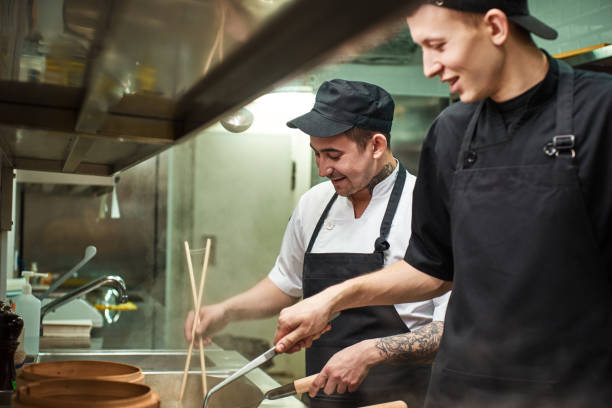 наслаждаюсь работой. два улыбающихся молодых помощника шеф-повара работают на кухне ресторана - главный курс стоковые фото и изображения