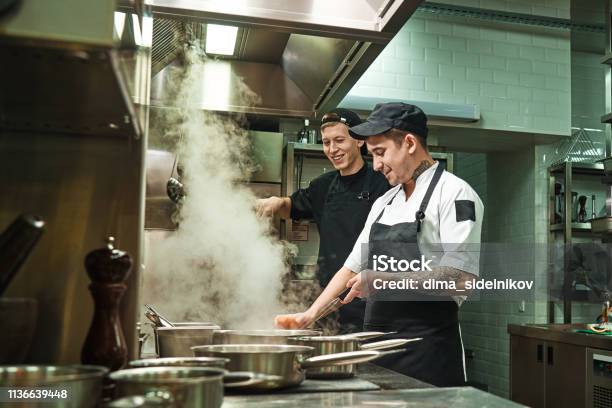 Positive Mood Side View Of Two Cheerful And Smiling Cooks In Uniform Are Preparing A Food In A Kitchen Restaurant Stock Photo - Download Image Now