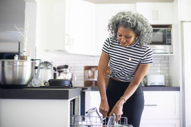 Mature Black Woman Loading Dishwasher A mature black woman with white curly hair loads empty dishes into the dishwasher for cleaning kitchen dishwasher stock pictures, royalty-free photos & images