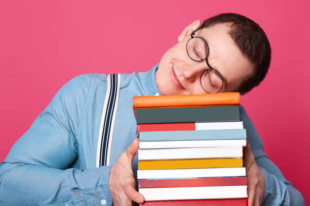 istock Handsome student wears blue shirt, suspenders and round glasses, puts his head on stack of books and sleeps. Tired guy prepares for exam for long time and feel exuasted, being glad falling asleep. 1136659636