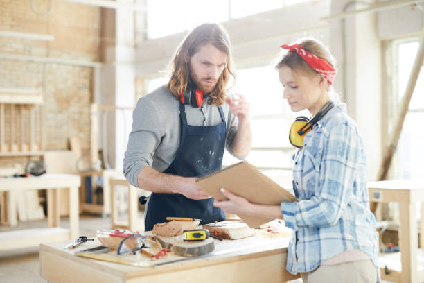 istock Serious thoughtful carpenters with ear protectors on necks standing in workshop and viewing sketch while talking about crafting wood detail 1136663917