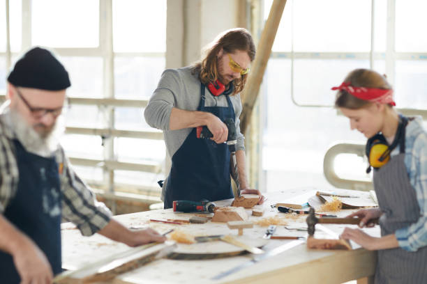 istock Serious concentrated handsome young carpenter in safety goggles standing at desk and using cordless drill while making hole in wooden piece 1136664063