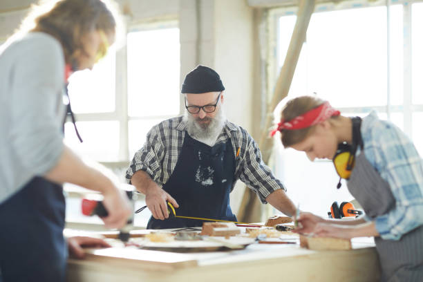 istock Serious gray-bearded workman concentrated on measuring standing at table and using self-retracting metal tape measure while working with wooden piece 1136664067
