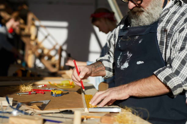 istock Close-up of concentrated senior carpenter with beard standing at table in workshop and drawing line on wooden plank 1136664115