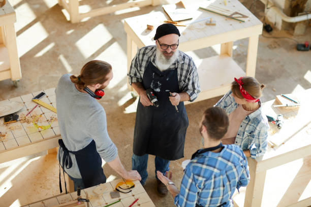 istock Carpentry workers chatting in workshop: serious senior carpenter in beanie hat holding drill bit and power tool while explaining young students how to use these tools 1136664242