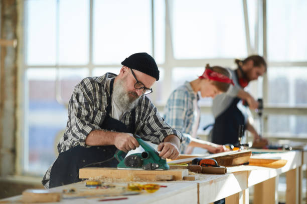 istock Serious busy senior bearded carpenter in apron and glasses standing at table and sanding wooden surface using power tool 1136664426