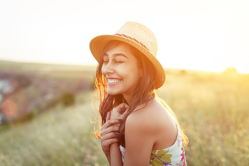 Side view of smiling happy woman in straw hat standing with eyes closed in nature and laughing in sunlight