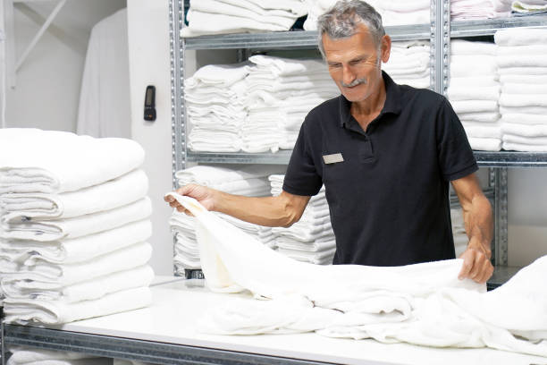 Elderly caucasian male laundry hotel worker folds a clean white towel. Hotel staff workers. Hotel linen cleaning services.