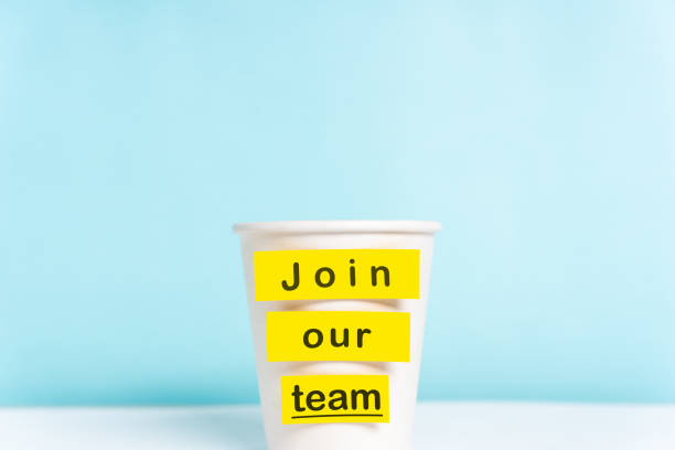 Concept of join our team or onboarding process. Paper cup with yellow top on desk with a paper cup over blue background. stock photo