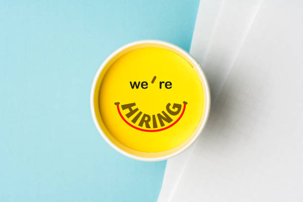 Concept of we're hiring or join our team or onboarding process. Paper cup with yellow top and text we're hiring illustrating a happy face emoticon, all this on a desk with blank documents over blue background. concepto de que estamos contratando o unirse a nuestro equipo o proceso de incorporación. taza de papel con tapa amarilla y texto que estamos contratando que ilustra un emoticono de cara feliz, todo esto en un escritorio con documentos en blanco sobre fon