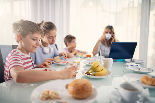 Three kids and mother eating breakfast in sunny living room. The mother is working from home.\nNikon D850