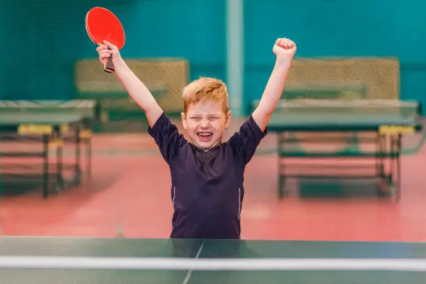 a boy in a gray t-shirt enjoys winning table tennis, blurred background, table tennis hall.