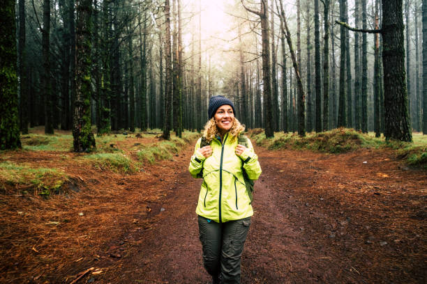 felice escursionista donna caucasica sorridere e godersi la natura camminando in una foresta con alberi alti - attività ricreativa all'aperto alternativa e stile di vita delle vacanze - sole in retroilluminazione e concetto di nebbia - tree area footpath hiking woods foto e immagini stock