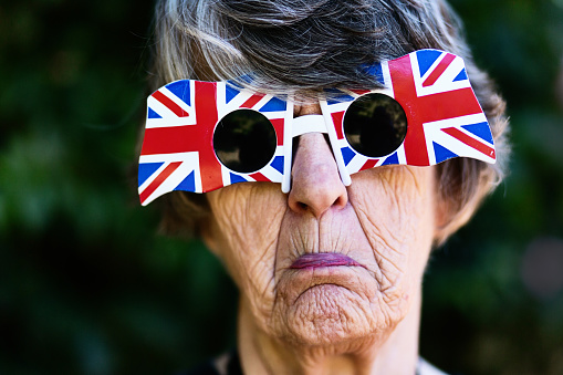 A senior female British patriot wears Union Jack sunglasses and frowns, very unhappy about Brexit.