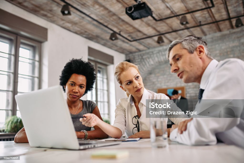 Multi-ethnic group of business people working on laptop during a meeting. Serious business team cooperating on a meeting while using laptop in the office. Business Stock Photo