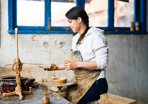 Mixed race woman molding clay on pottery wheel and using smart phone with dirty hands.