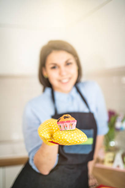 Young woman hold muffin on foreground with cooking gloves.