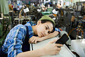Sad attractive young woman worker in beanie hat being bored at workplace, she using smartphone and leaning on industrial equipment