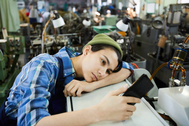 Sad attractive young woman worker in beanie hat being bored at workplace, she using smartphone and leaning on industrial equipment stock photo