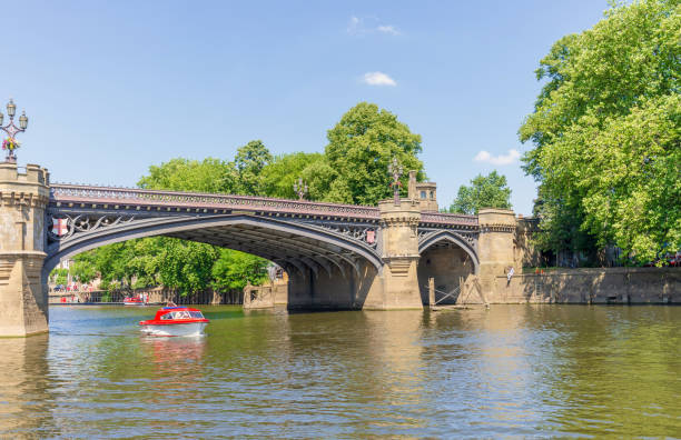 A small boat passes under a bridge. The 19th Century Skeldergte Bridge in York.  A small boat passes underneath while trees line the river bank and a blue summer sky is above. york yorkshire stock pictures, royalty-free photos & images