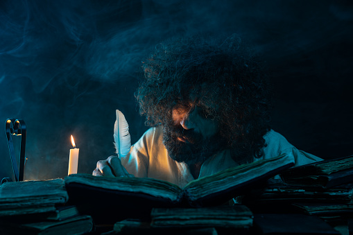 Portrait of mature adult man in medieval costume writing with quills pen in dark. He has long hair and beard. The candlelight is on the left side of frame. Shot with a full frame mirrorless digital camera.