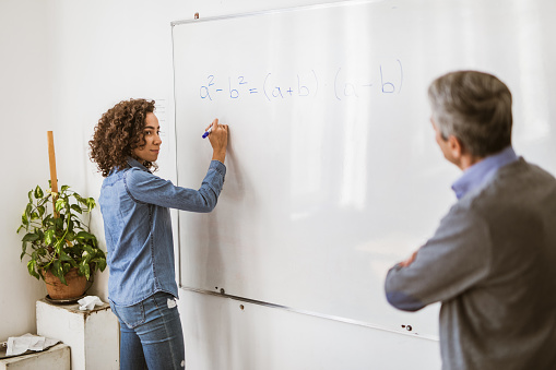 African American female student writing a formula on whiteboard while her teacher is observing it in the classroom.