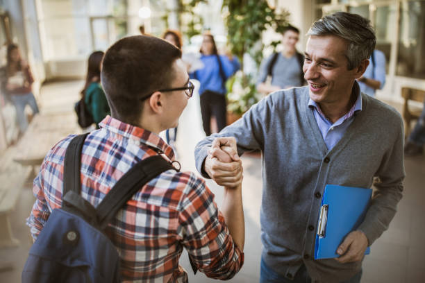 Happy teacher and high school student greeting in a hallway. Happy mid adult teacher and male student greeting in a school hallway. There are people in the background. teacher reprimanding student in hallway stock pictures, royalty-free photos & images