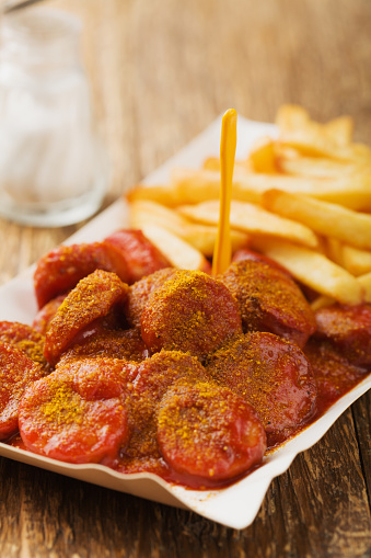 Traditional German currywurst, served with chips on disposable paper tray. Wooden table as  background.