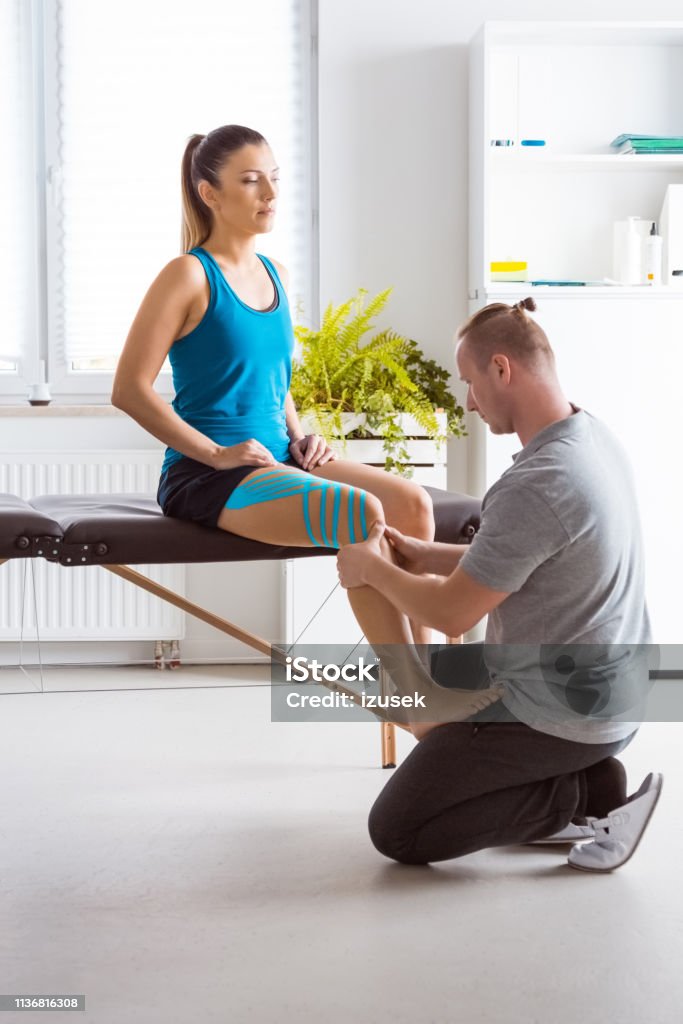 Physiotherapist massaging young woman's leg Physical therapist giving leg massage to young woman. Patient having elastic therapeutic tape on her knee. Recovery Stock Photo