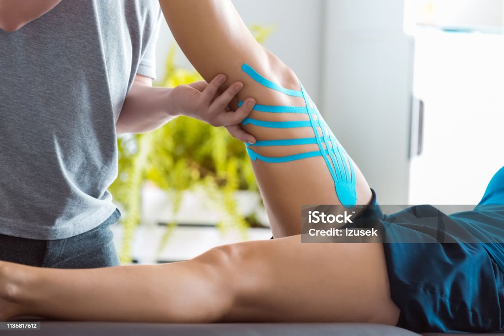 Physiotherapist stretching young woman's leg Physical therapist giving leg massage to young woman. Patient having elastic therapeutic tape on her knee. Close up, unrecognizable people. Sports Medicine Stock Photo