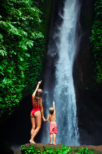 In tropical jungle happy mother with kid stand under waterfall, explore rainforest nature. Travel adventure, hiking activity with child. Lifestyle on family summer vacation and weekend walking tour.